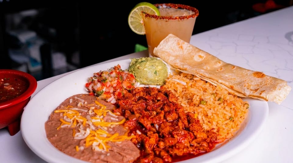 A Mexican platter with rice, beans, meat, guacamole, salsa, tortillas, and a margarita.