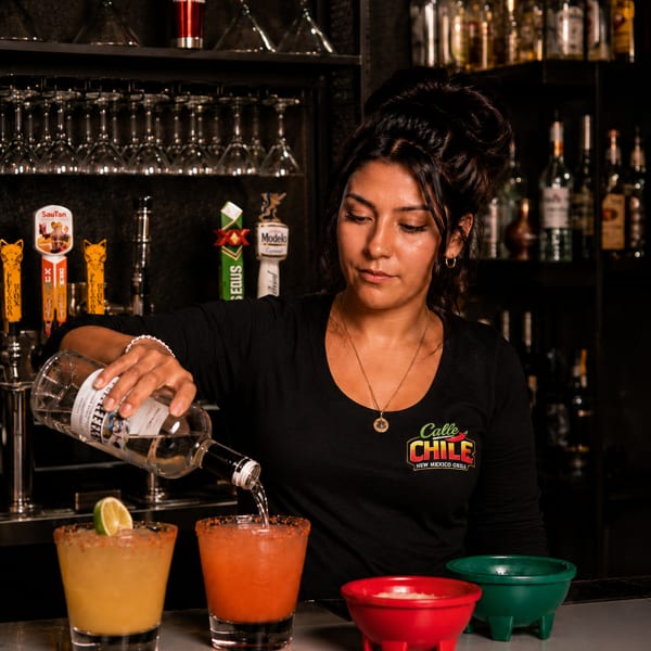 A bartender pouring a drink into a glass with two colorful margaritas on a bar.