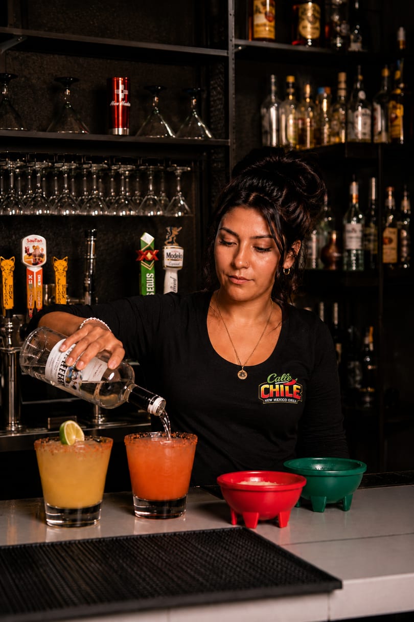 A bartender pouring a drink into a glass with two colorful margaritas on a bar.
